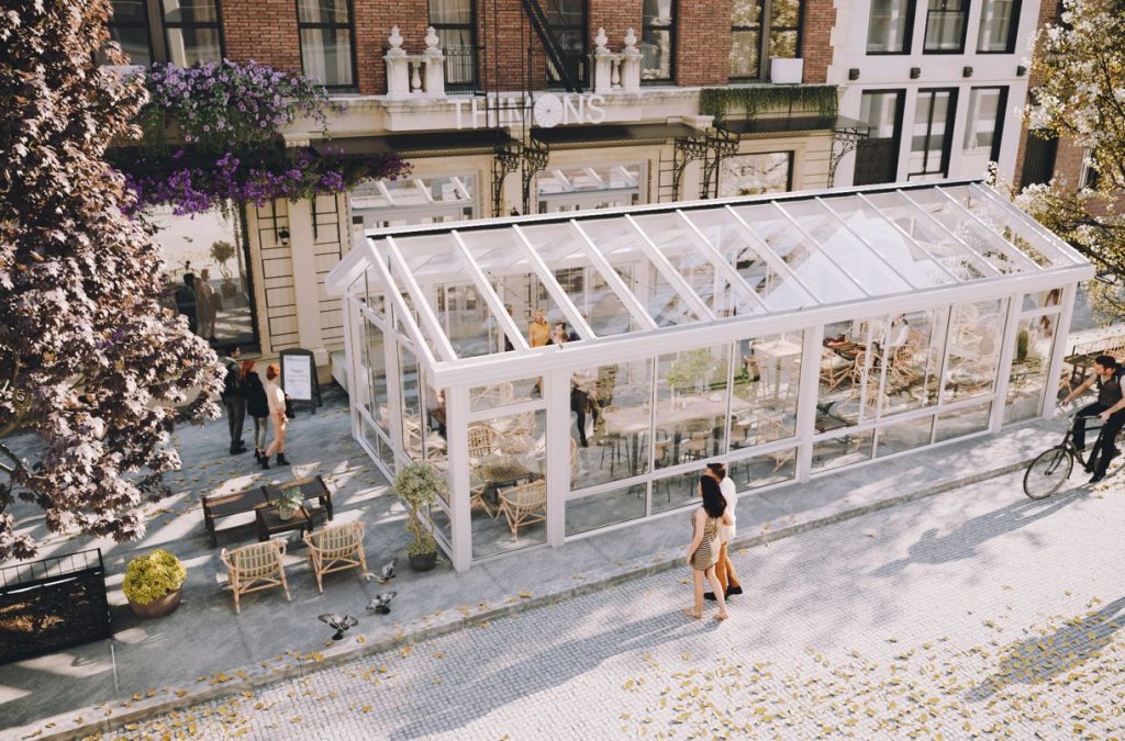 Ett stort fristående uterum Exal Nordic 140 med sadeltak som används som uteservering till en restaurang. Rummet har ett sadeltak och vita aluminiumstommar och ligger på en gågata där folk promenerar och cyklar förbi. Ett tegelhus syns i bakgrunden och blommande träd vid sidorna om rummet.