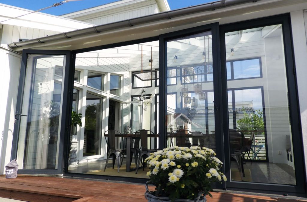 Ett smakfullt expodul uterum vinterträdgård med praktiska vikpartier som vätter ut mot en väl inredd uteplats. Solen tittar in via det isolerade glastaket och speglar sig mot husväggen.